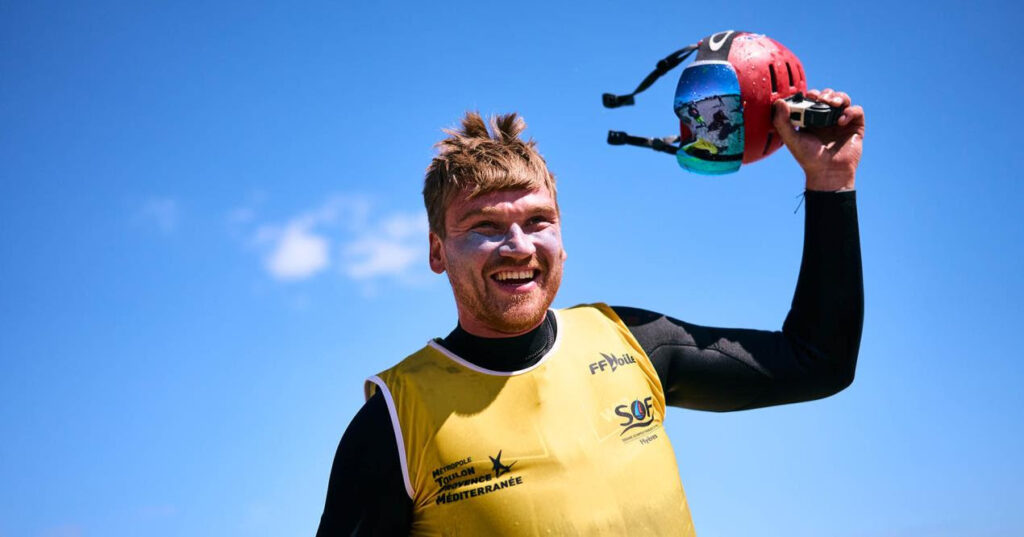 Connor Bainbridge  holding his hat, in celebration. He is smiling and looking very pleased. His hair is slightly messy from wearing his protective helmet.